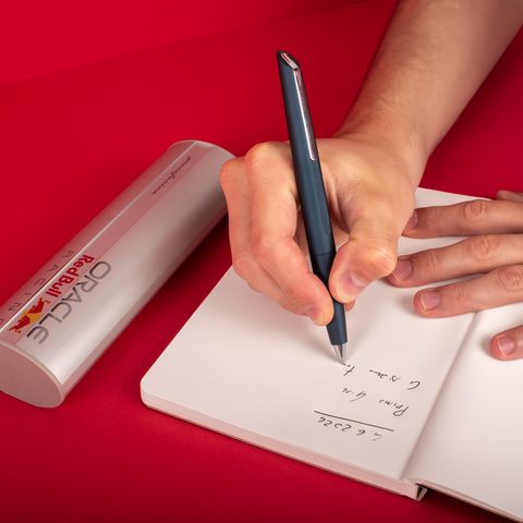 Person writing in a stone paper notebook with the Pininfarina Segno "PF TWO Oracle Red Bull Racing" in its ballpoint version, next to its packaging tube.