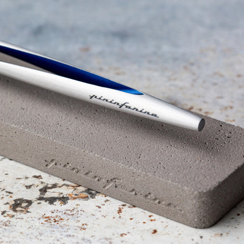 Close up of the Pininfarina Segno "Aero" inkless pen with ethergraf tip with aluminum silver and blue body in its concrete base in a rusty white table.