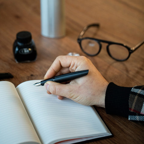 Hand writing in a notebook with the Pininfarina Segno "PF TWO" in its black fountain pen version in a desk with a bottle with ink and glasses.