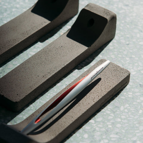 Pininfarina Segno "Aero" inkless pen with ethergraf tip with aluminum silver and red body in its concrete base and other concrete bases on a table.