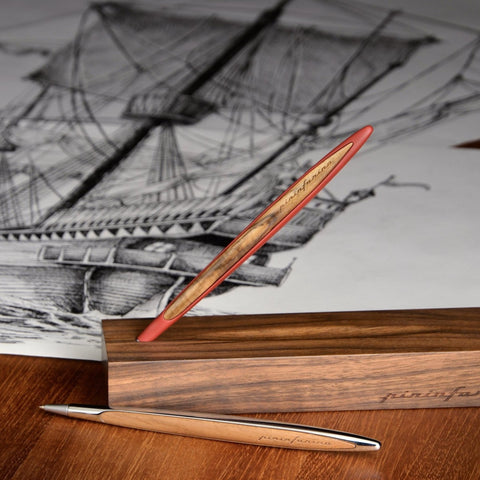 Pininfarina Segno "Cambiano Ink" ballpoint pen available in, red, blue and silver aluminum body and walnut wood inserts standing in its base in a desk with drawings.
