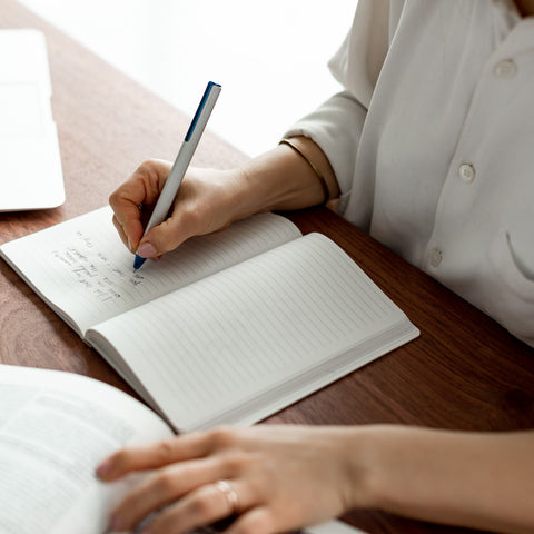 Woman writing in a stone paper notebook with the Pininfarina Segno "PF ONE Ballpoint" pen in its bicolor version; white body and blue tip and clip. 