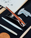 FO - Forever Made In Italy "Piuma Leonardo Special Edition" ethergraf inkless or ballpoint pen in its pear wood steam version, standing in its stainless steel ondulated base, next to other piumas and rulers. 