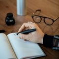 Hand writing in a notebook with the Pininfarina Segno "PF TWO" in its black fountain pen version in a desk with a bottle with ink and glasses.