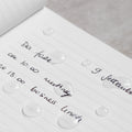 Close up of the Pininfarina Segno "Stone Paper Notebook" in its ruled version with notes written and drops of water, showing that the water doesn't absorb and it's waterproof.
