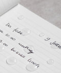 Close up of the Pininfarina Segno "Stone Paper Notebook" in its ruled version with notes written and drops of water, showing that the water doesn't absorb and it's waterproof.