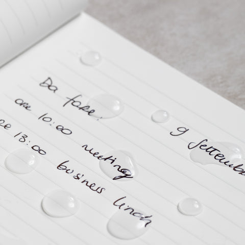Close up of the Pininfarina Segno "Stone Paper Notebook" in its ruled version with notes written and drops of water, showing that the water doesn't absorb and it's waterproof.