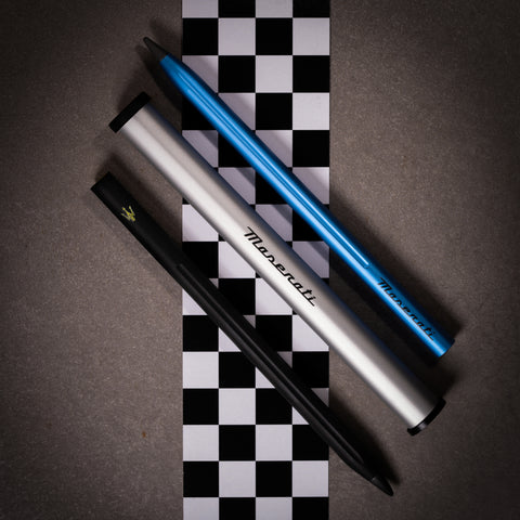 Pininfarina Segno "SMART Maserati Edition" pencil with grafeex tip in its black and blue versions next to its packaging tube in a checkered flag and concrete background.